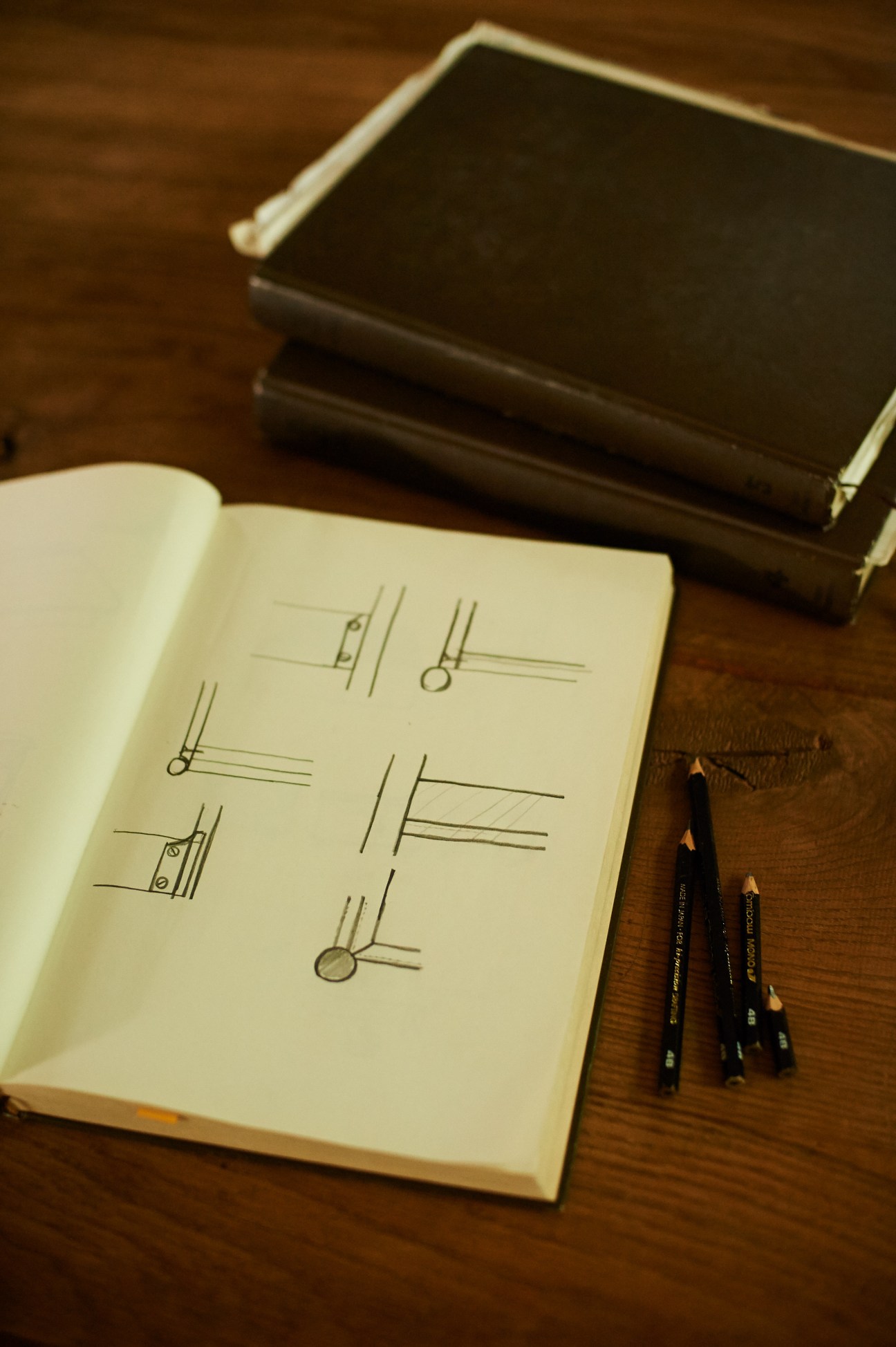 Open hardcover notebook with furniture design sketches and pencils on wooden surface.