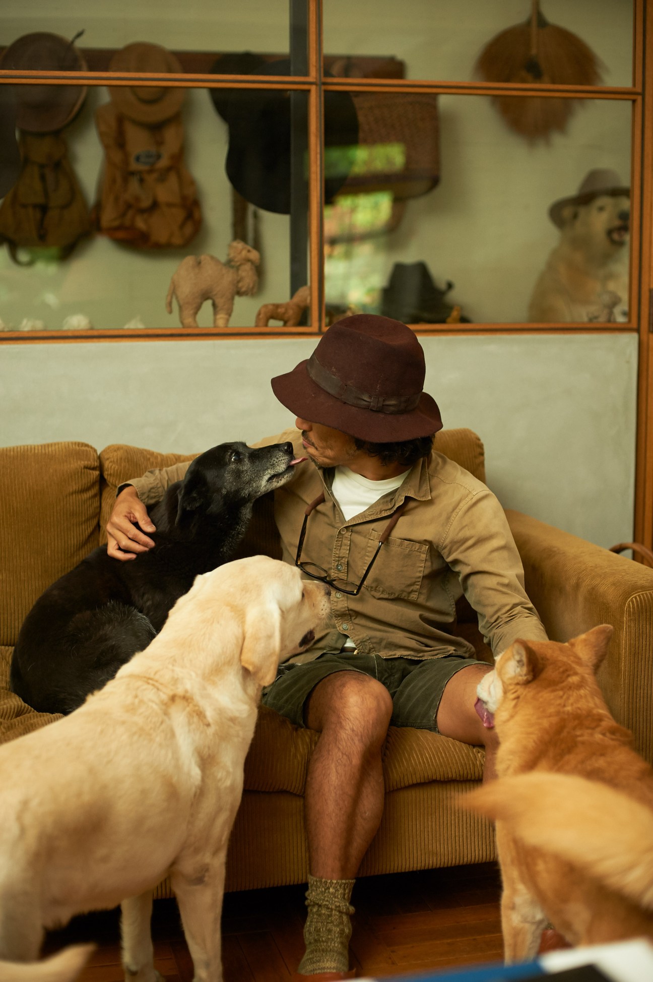 Man wearing brown hat sitting on corduroy sofa with three dogs around him.