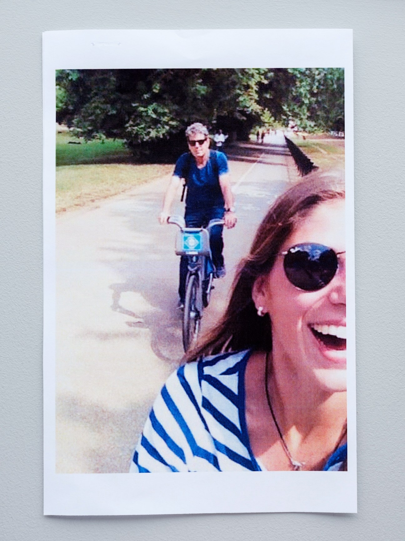 Two people on a bike path, one riding a Bixi bicycle in the background and another smiling at the camera in the foreground wearing sunglasses and striped clothing.