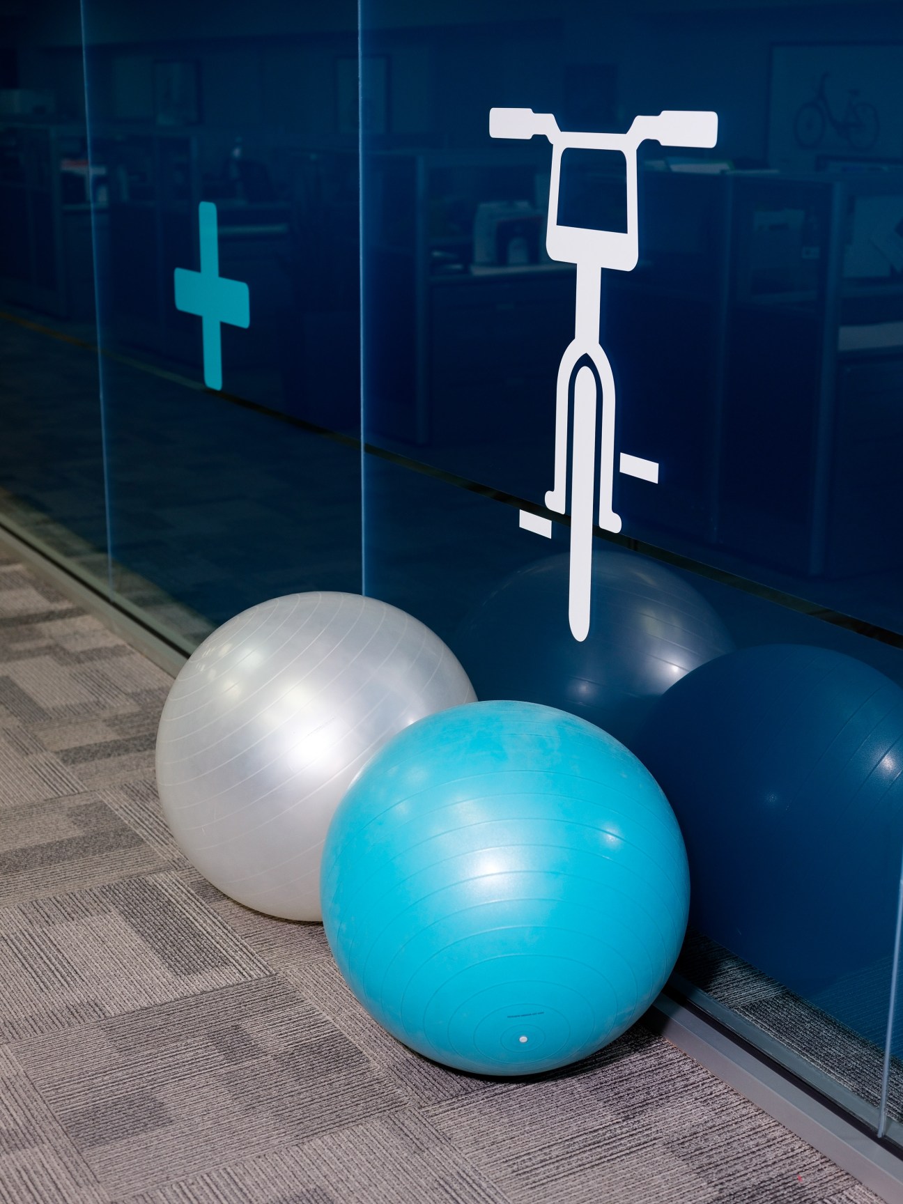 Exercise balls in turquoise and white sit on carpet next to a blue glass wall with white bike and medical cross icons.