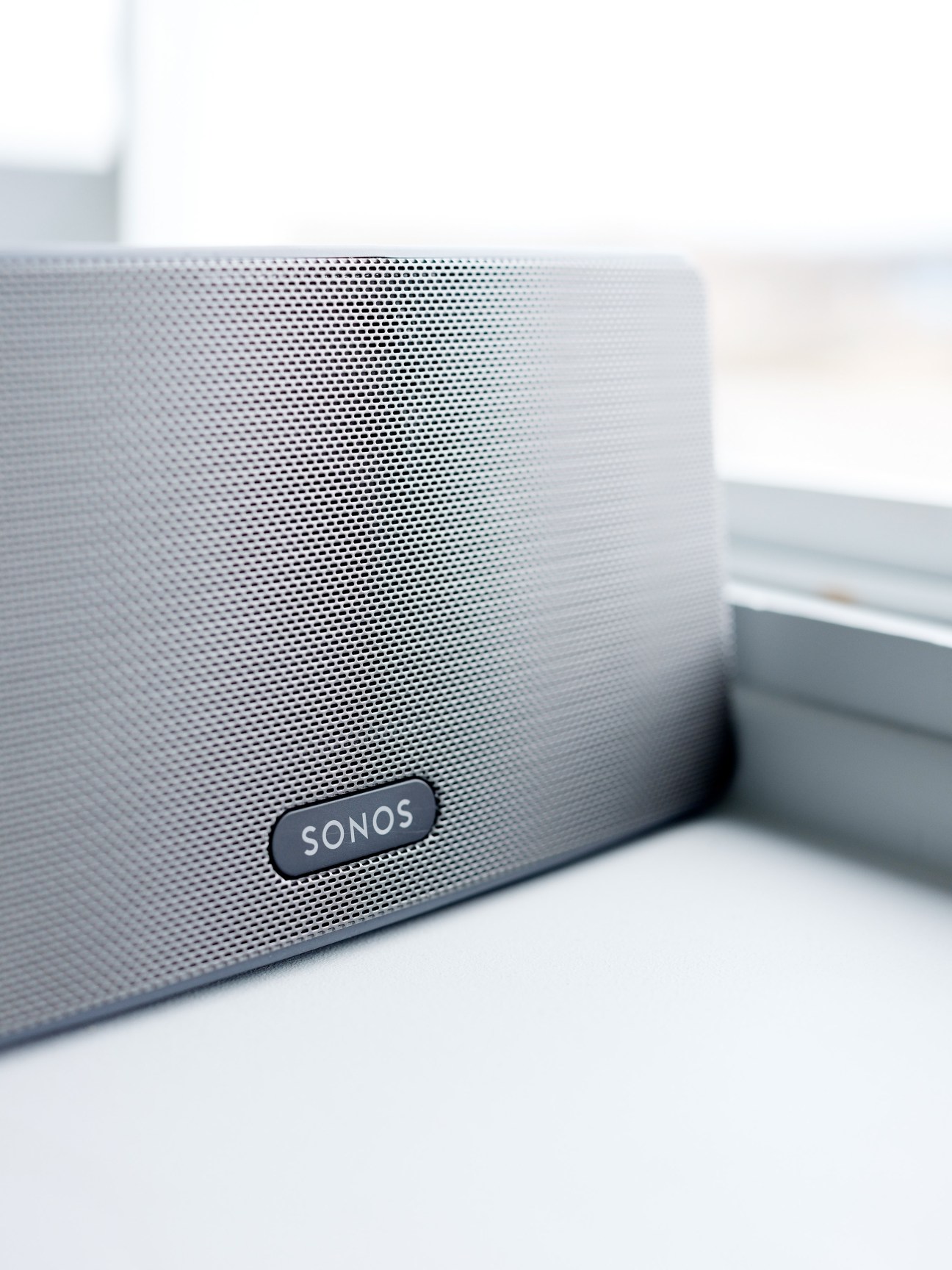 A white Sonos speaker with perforated mesh grille and dark oval logo badge on a light surface.