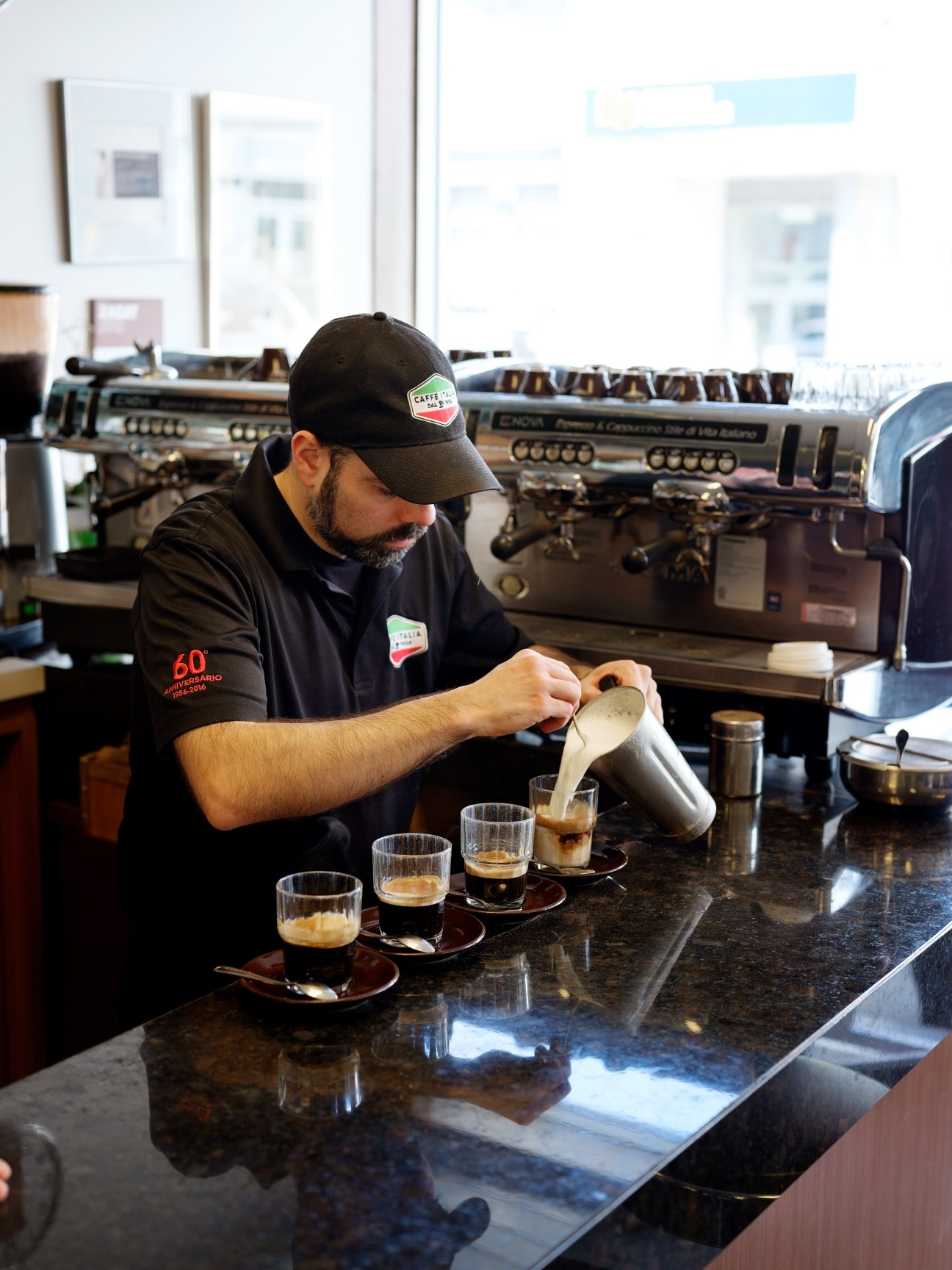 Barista pouring milk into espresso cups at a café counter with professional espresso machine in background.