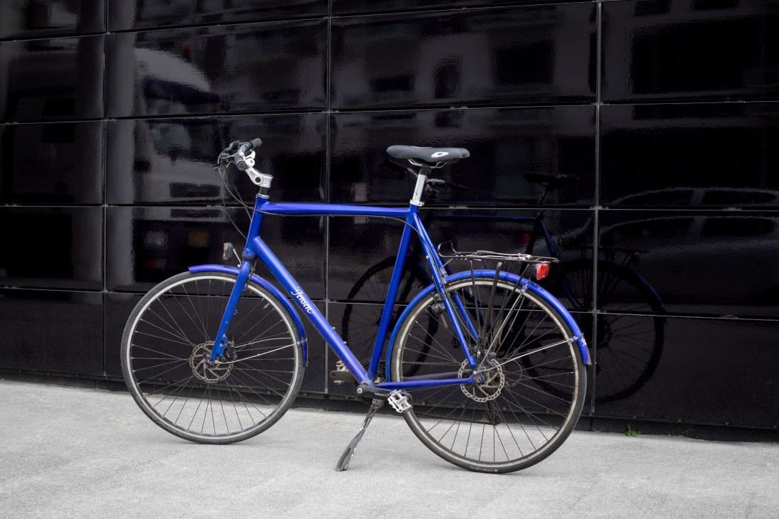 A blue bicycle with disc brakes leaning against a dark tiled building wall.
