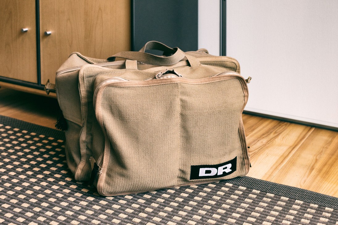 Beige canvas DR bag with dark strap and black logo label on wooden floor.