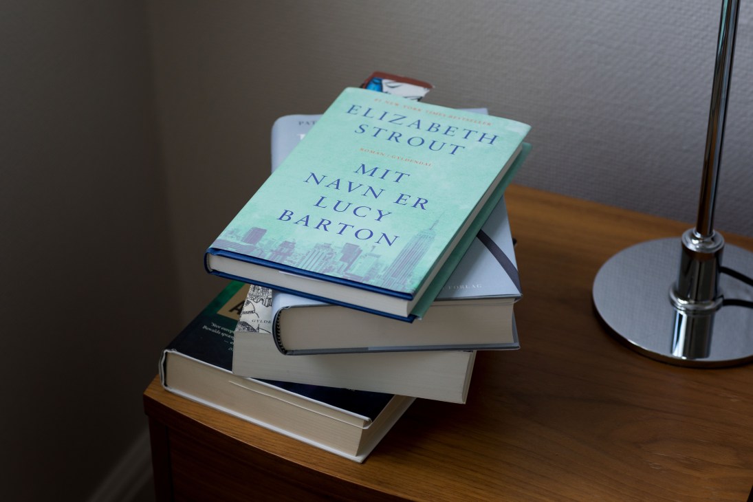 Stack of books on a wooden desk including Elizabeth Strout's 'Mit navn er Lucy Barton', next to a desk lamp.
