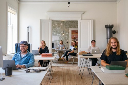 Open office workspace with wooden flooring, multiple staff members at desks, and wooden structural elements creating meeting pods.