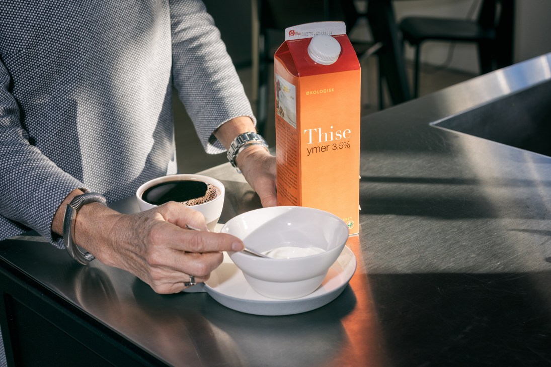 Woman's hands holding a white bowl with ymer yogurt and a carton of Thise brand ymer on a table.