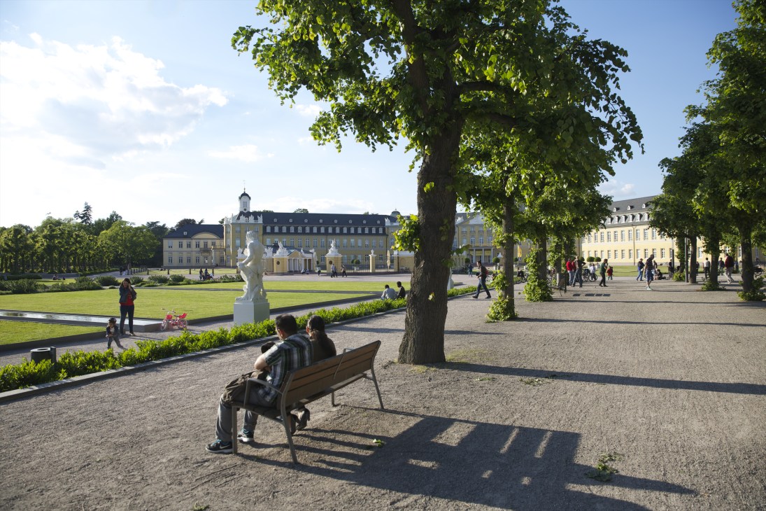 Karlsruhe Palace with radiating tree-lined avenues and people relaxing on benches in the palace gardens.