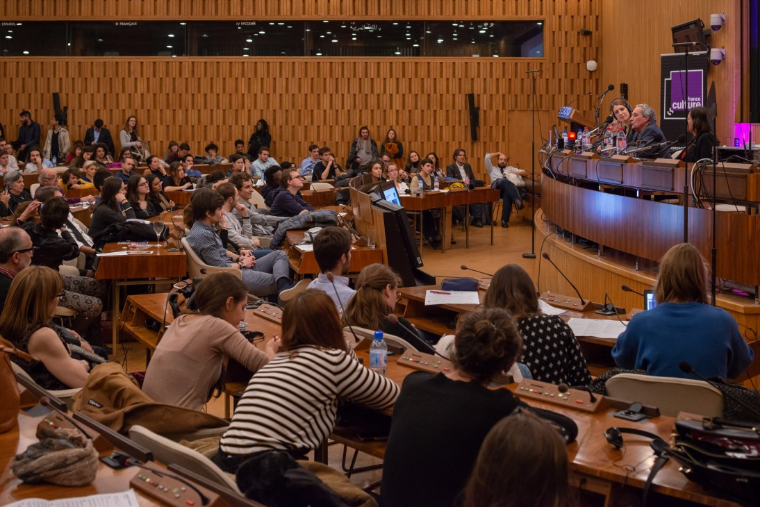 A crowded lecture hall with seated audience members facing a panel of speakers at a table with microphones and water glasses.
