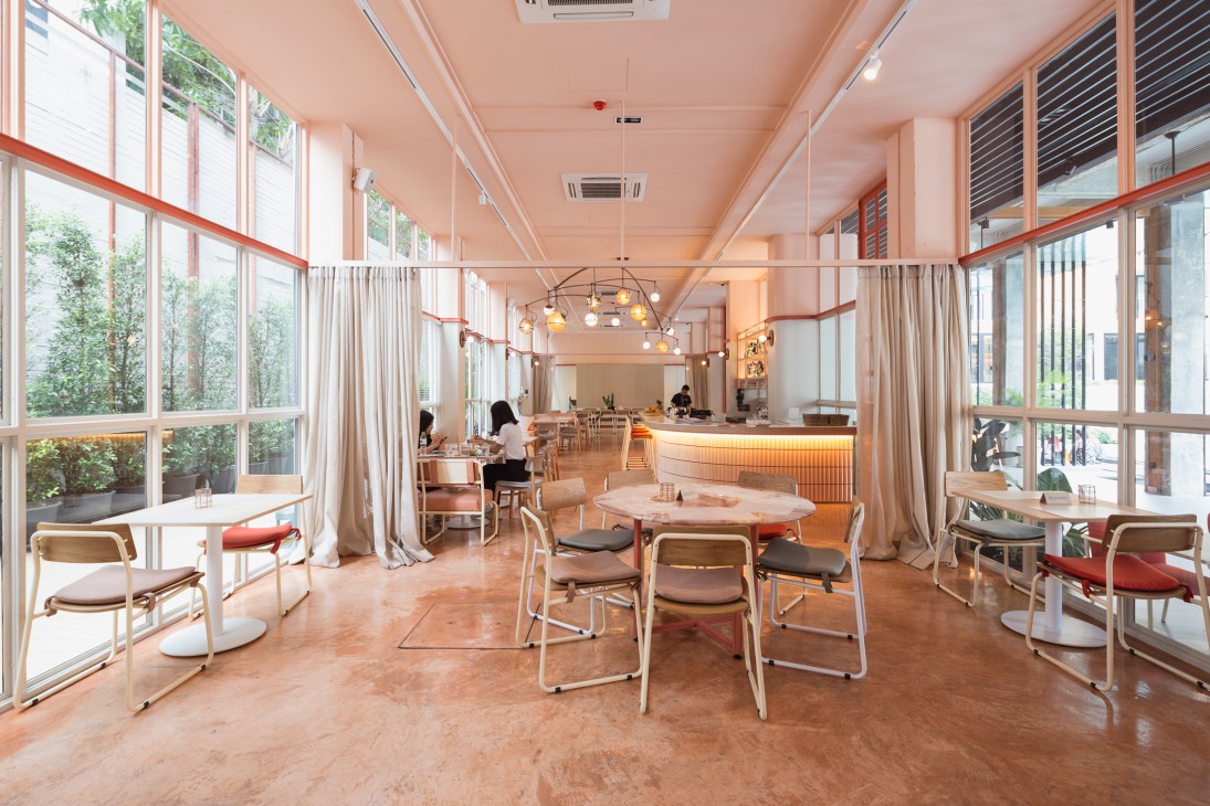 Modern hotel restaurant and lounge with floor-to-ceiling windows, coral pink ceiling, wooden furniture, and pendant lighting fixtures.