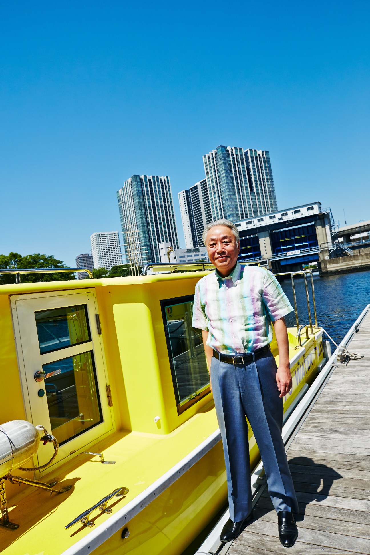Hajime Tabata stands on a dock next to a bright yellow water taxi with Tokyo's skyline in the background.