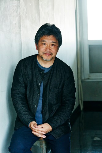 Japanese film director Hirokazu Koreeda sitting against a white wall wearing a dark jacket.