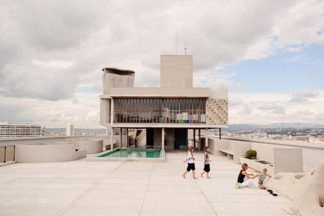 Children play on the rooftop terrace of Le Corbusier's Cité Radieuse with a swimming pool and concrete brutalist architecture.