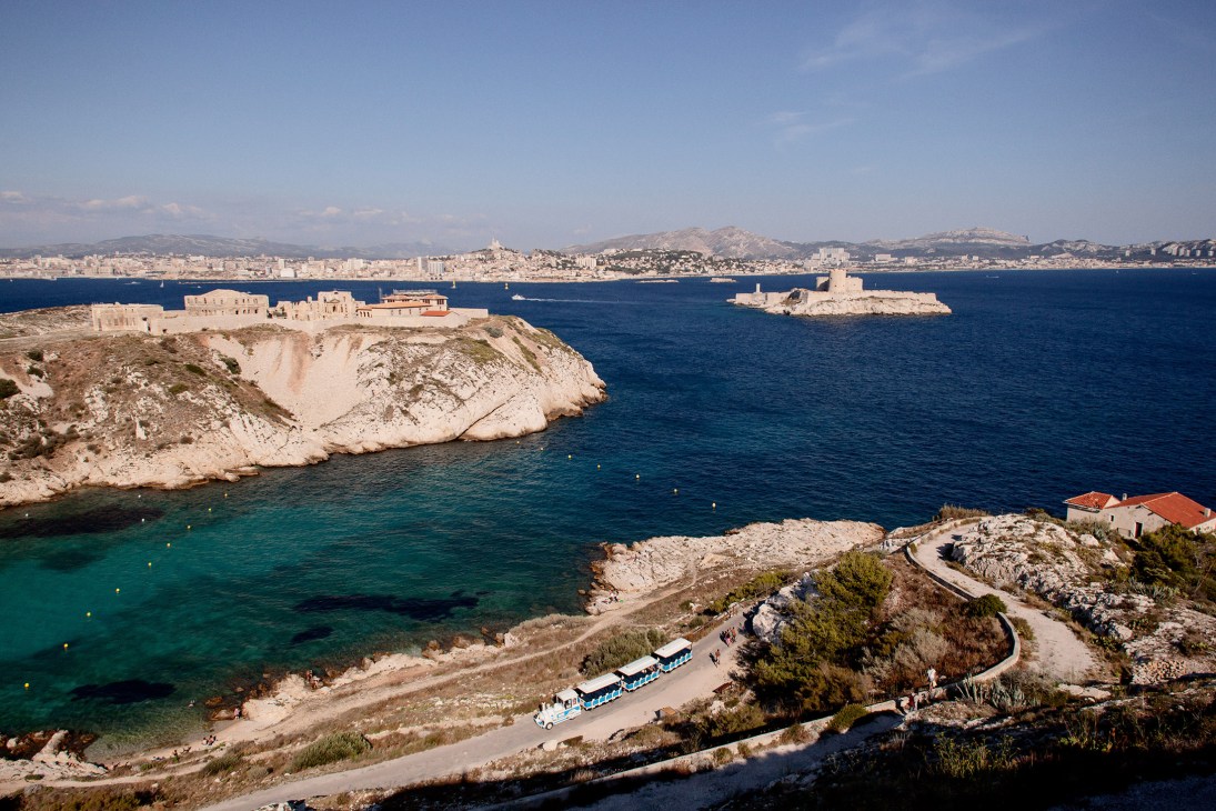 Château d'If fortress on a rocky island in the Mediterranean Sea with Marseille cityscape and mountains in the background.