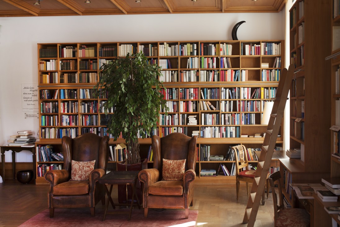 Library interior at Schloss Elmau with wooden bookshelves, leather armchairs, and a crescent moon sculpture on the wall.