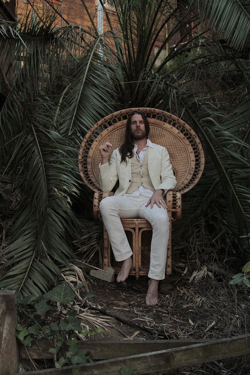 Man with long dark hair in cream suit seated on wicker peacock chair surrounded by tropical plants.