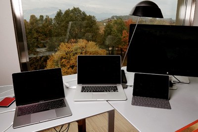 Multiple Apple devices including laptops and tablets arranged on a desk with a landscape view behind.