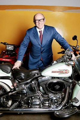 Man in blue blazer and glasses posing with a Harley Davidson motorcycle against a yellow wall.