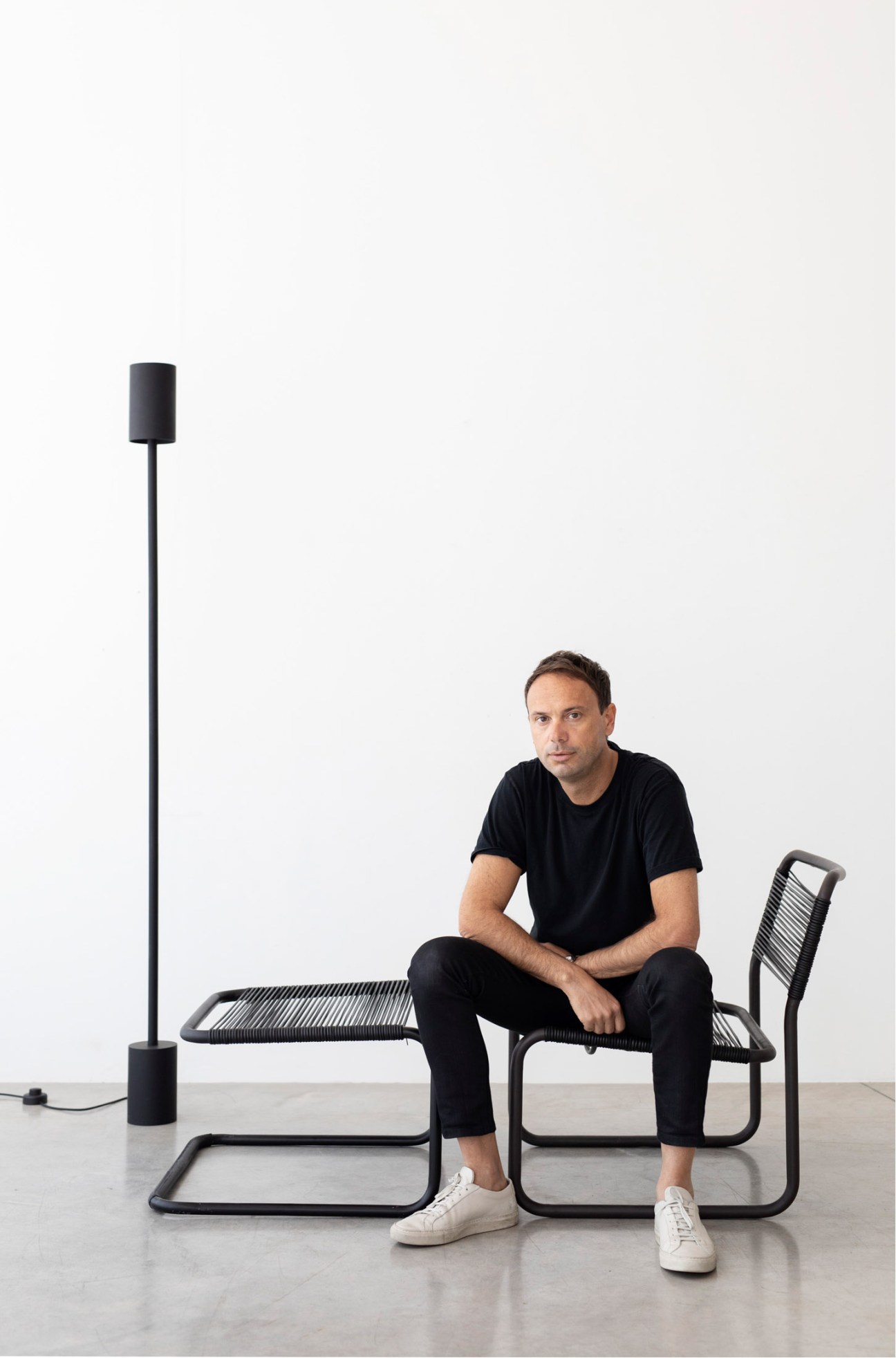 Man in black clothing sitting on a modern chair with metal frame and rope seat, next to a floor lamp.