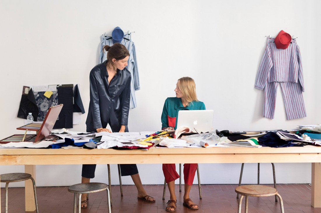 Two women work at a design table surrounded by fabric swatches and clothing samples displayed on wall-mounted stands.