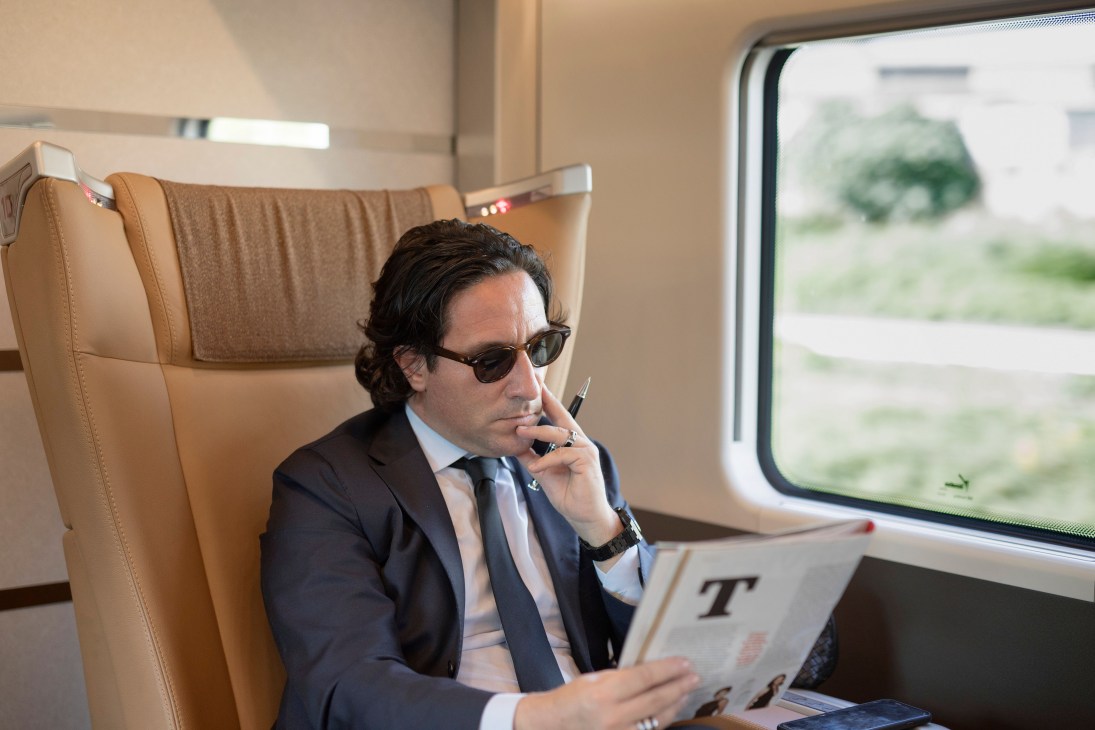 Man in business suit reading newspaper on Frecciarossa train with reclined leather seat.