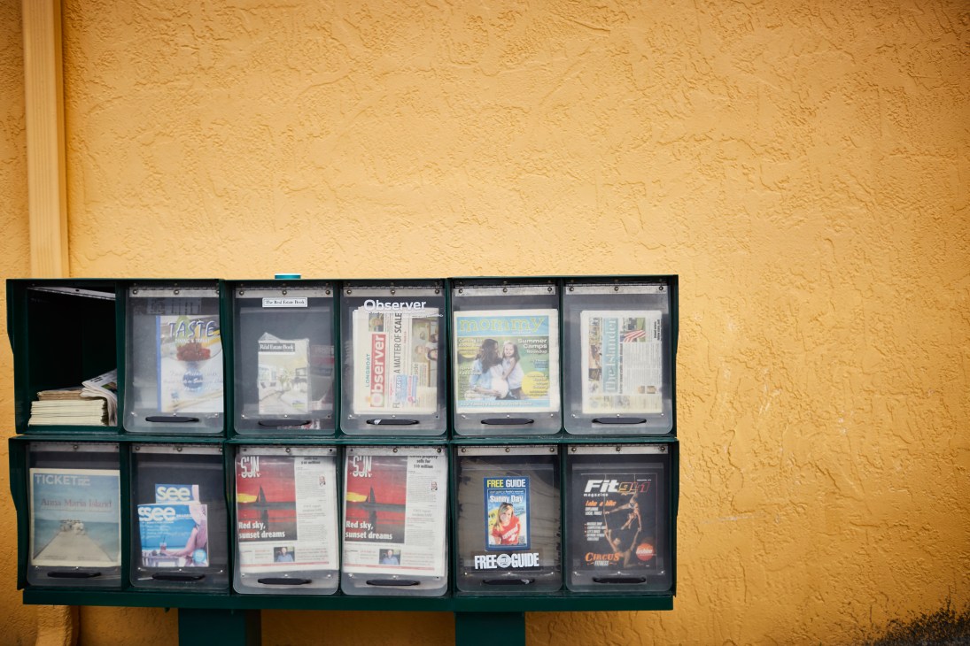 Newspaper and magazine stand mounted on yellow wall displaying various publications including The Islander.