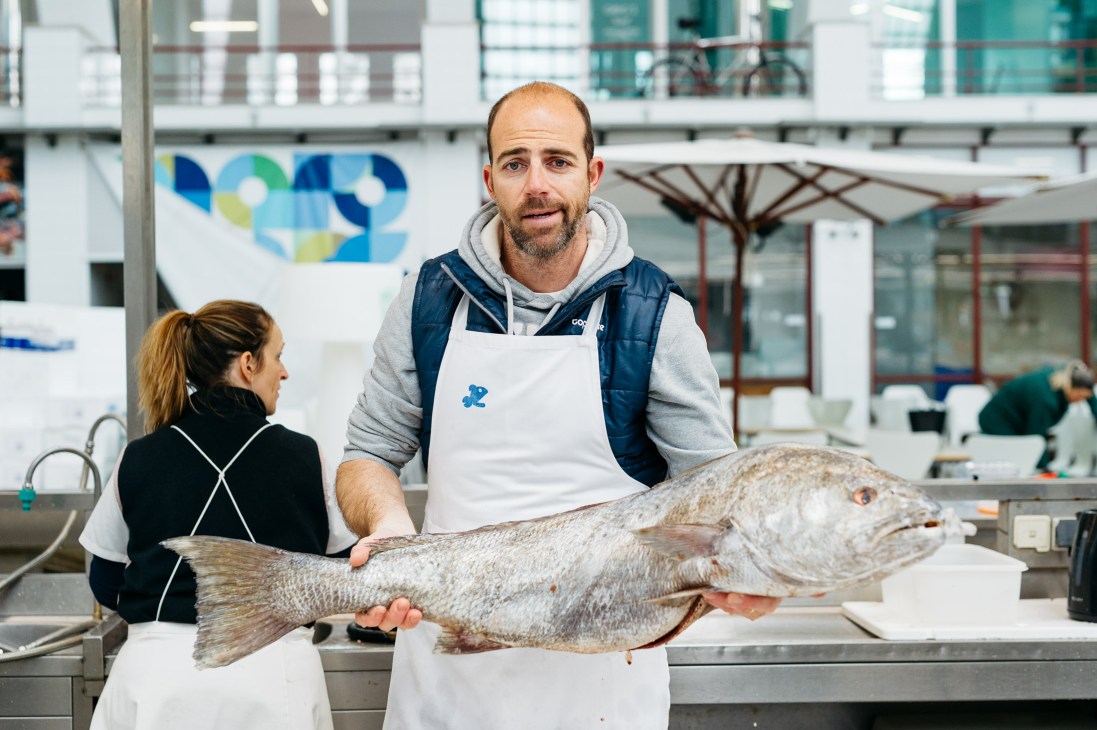 A man in a white apron and blue vest holds a large fresh fish while speaking with a woman in a kitchen facility.