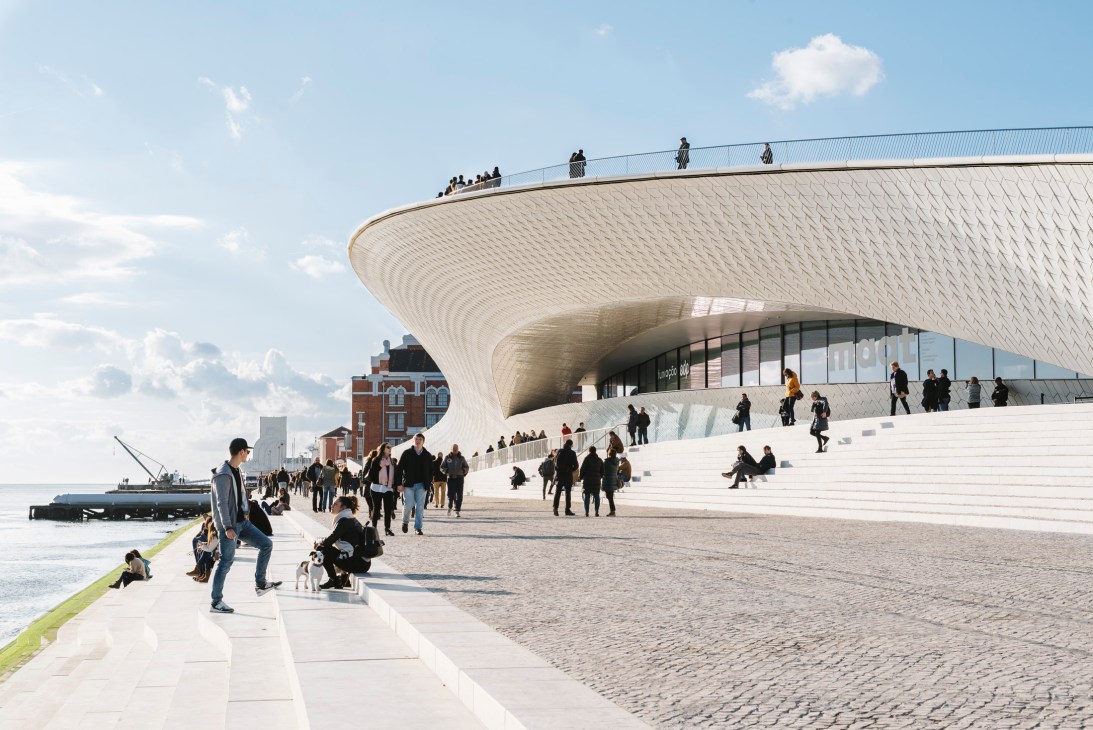 Modern waterfront building with curved white architecture and people on plaza steps overlooking water.