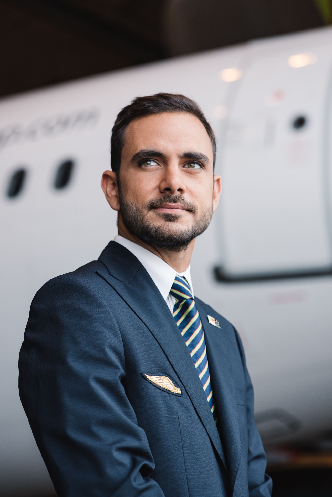 Male flight attendant in TAP Portugal uniform with blue blazer and striped tie smiling at camera in front of aircraft.
