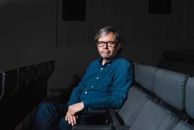 Pedro Borges, a man wearing glasses and a blue shirt, sits on a grey couch in a dark room.