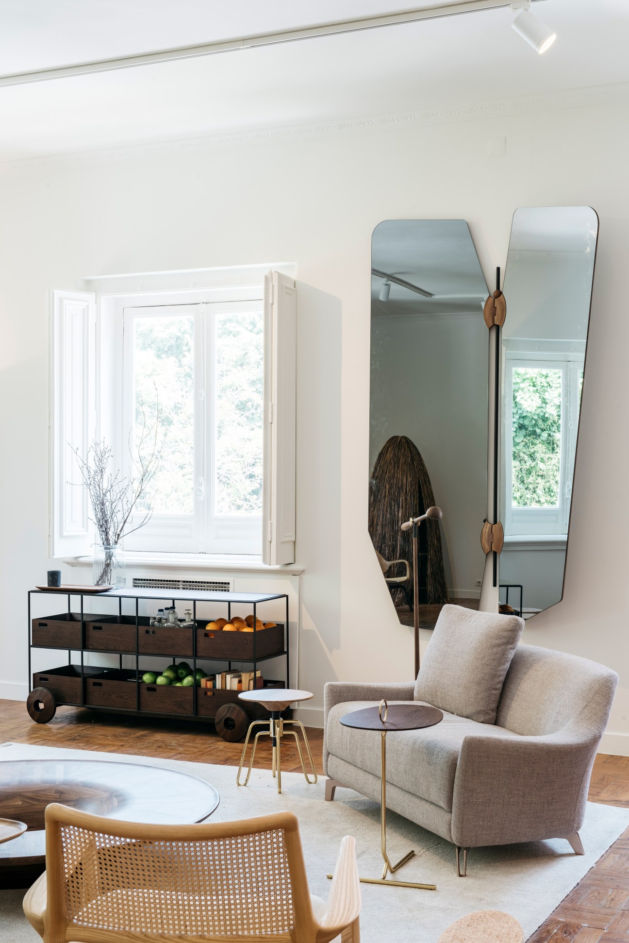 Modern interior space with beige armchair, tall mirror, wooden storage cart, and natural light from window.