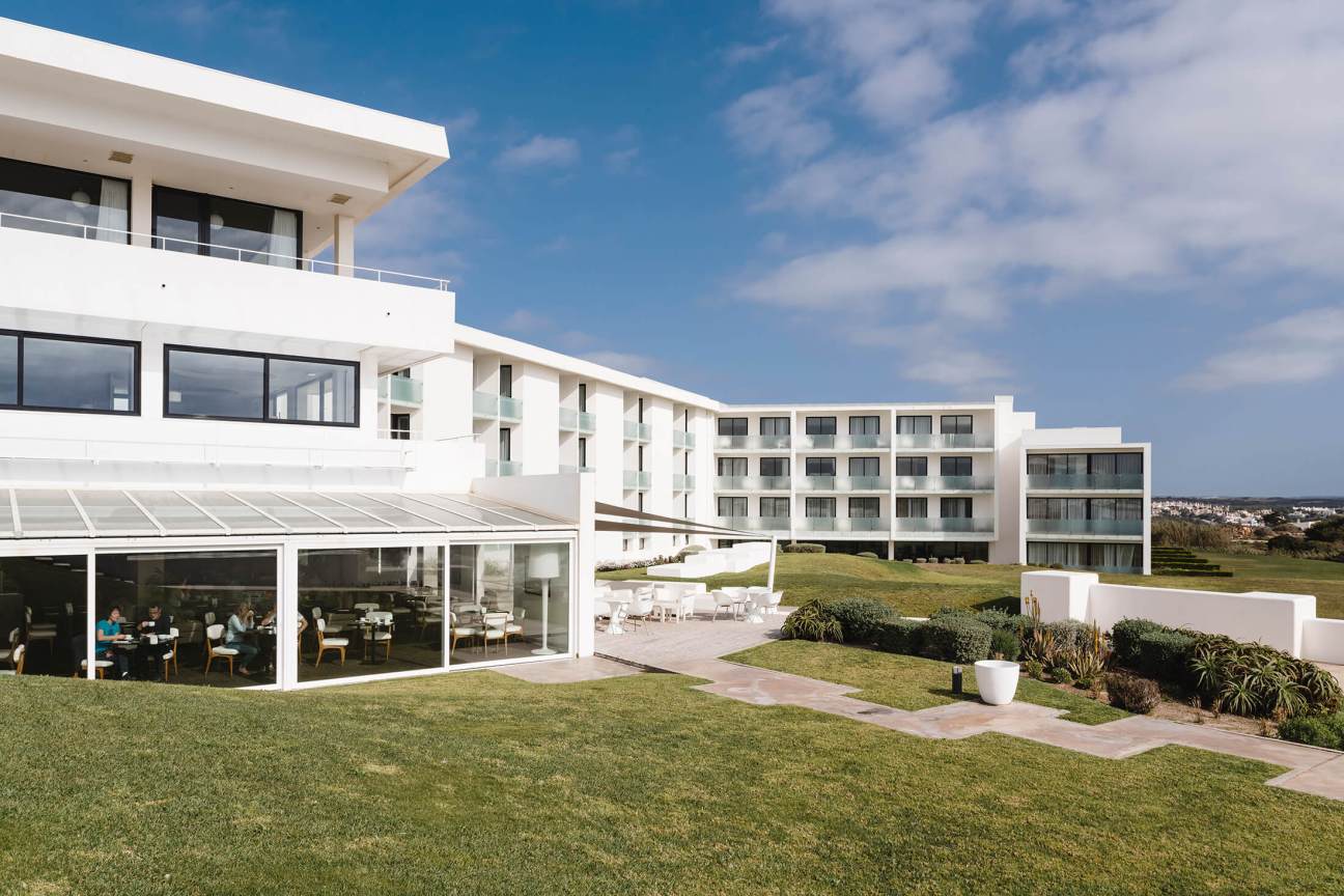 Modern white hotel building with curved architecture overlooking green lawns and ocean under blue sky.