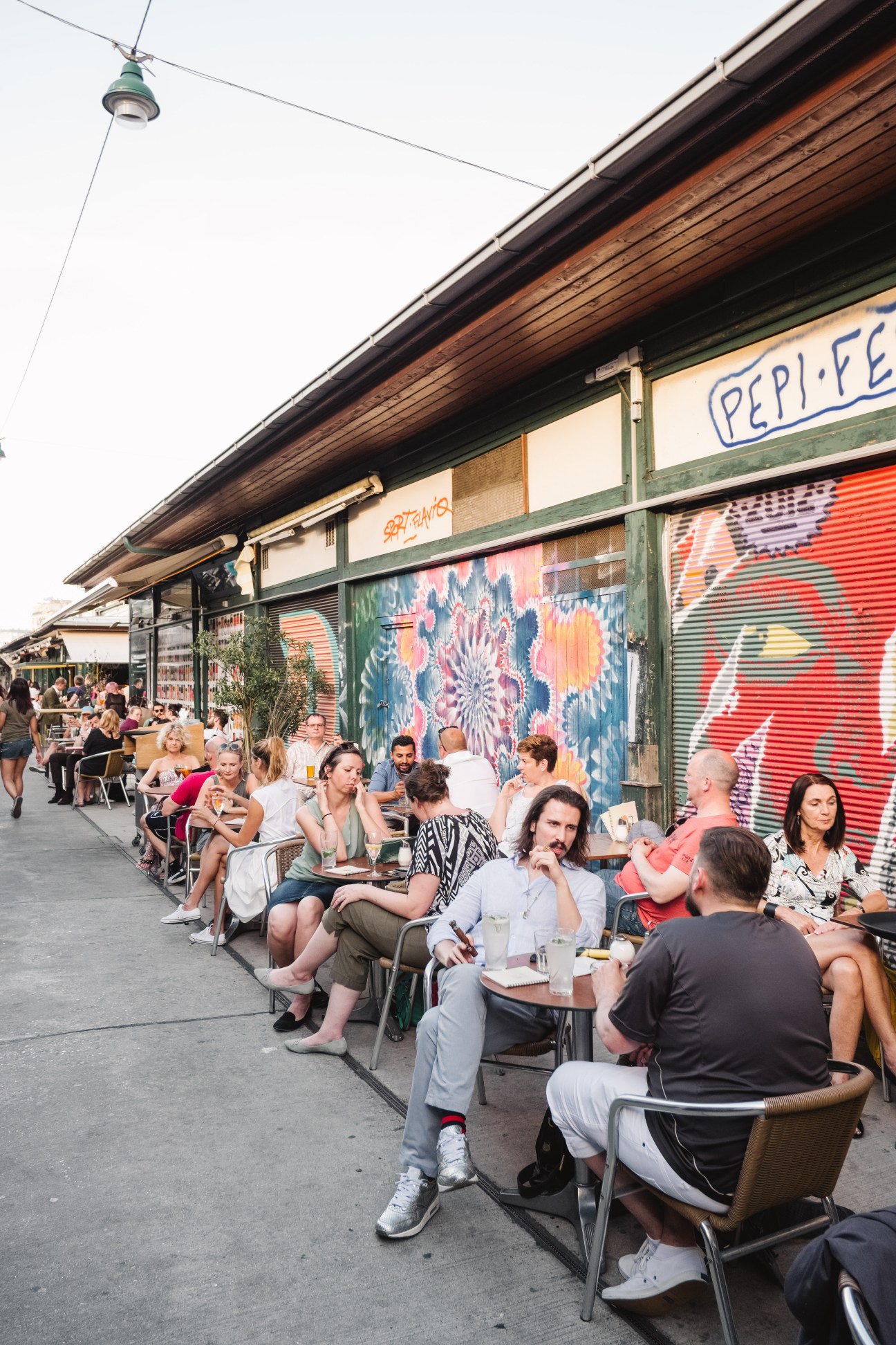 Outdoor dining scene with people seated at tables along a street with colorful street art on shutters