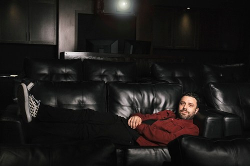 Man in red shirt lounging on black leather sofa in dimly lit room.