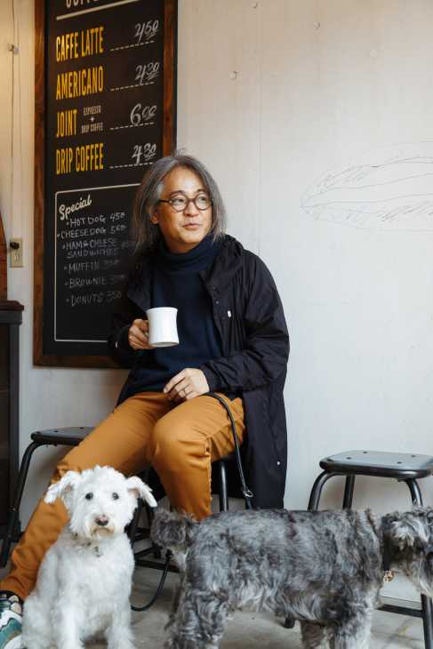 Person holding coffee cup at Little Nap Coffee Roasters café with dogs nearby and menu board in background.