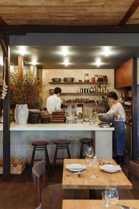 Modern dining space with open kitchen counter where two chefs work, shelves displaying cookware and ingredients, wooden beams overhead.