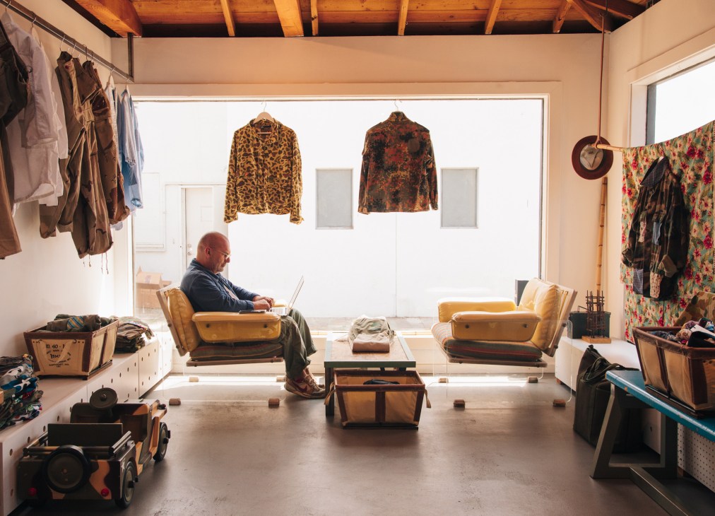 Man working on a laptop in a studio filled with vintage clothing, textiles, and accessories displayed on racks and shelves.