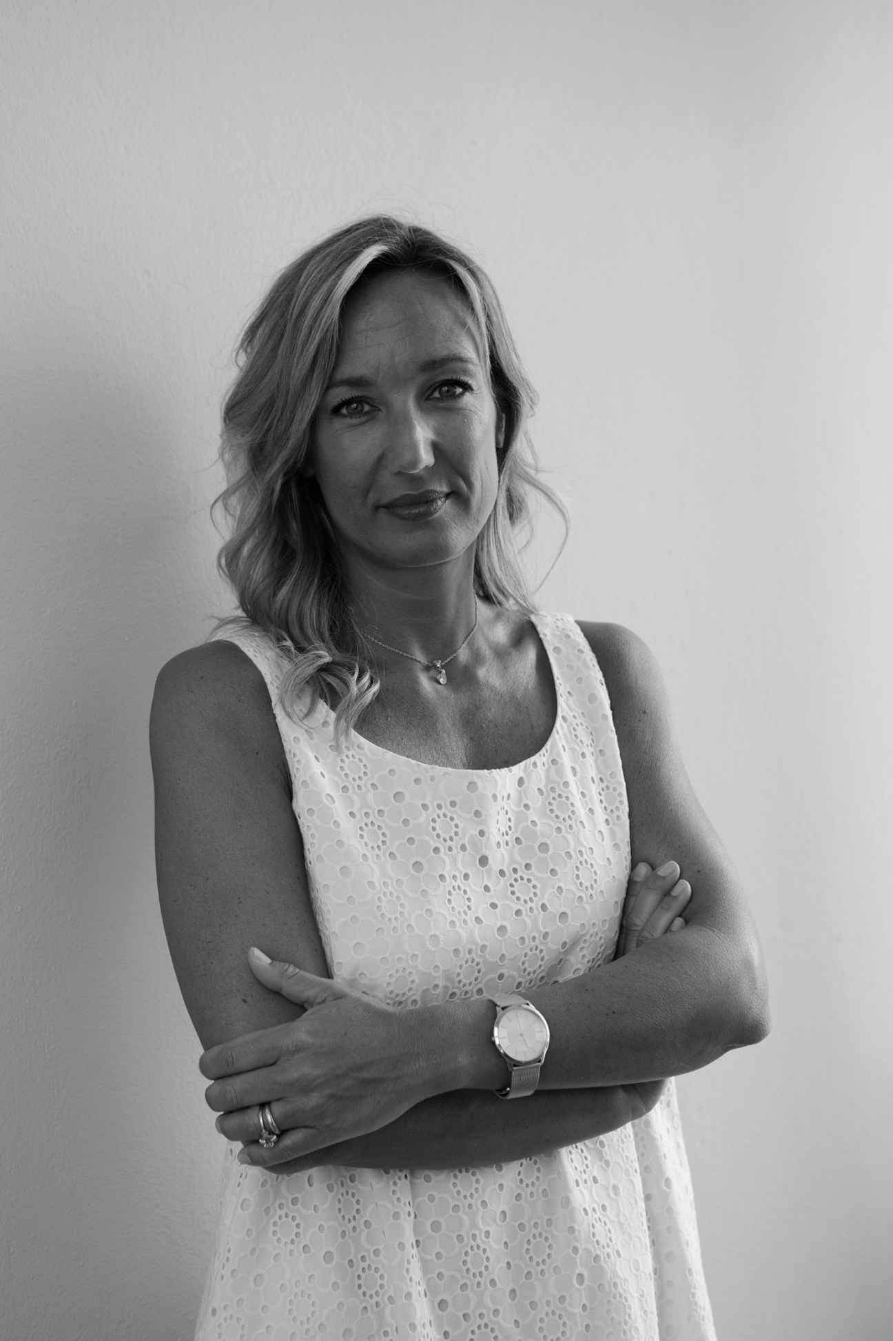 Black and white portrait of a woman in a white eyelet dress wearing a watch, standing with arms crossed.