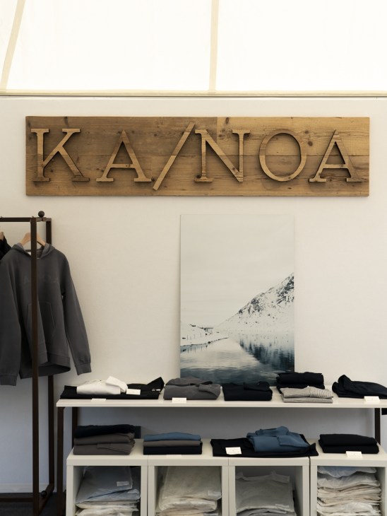 Ka/Noa store interior in Zürich with wooden signage, clothing displayed on shelves, and a snowy mountain photograph on the wall.