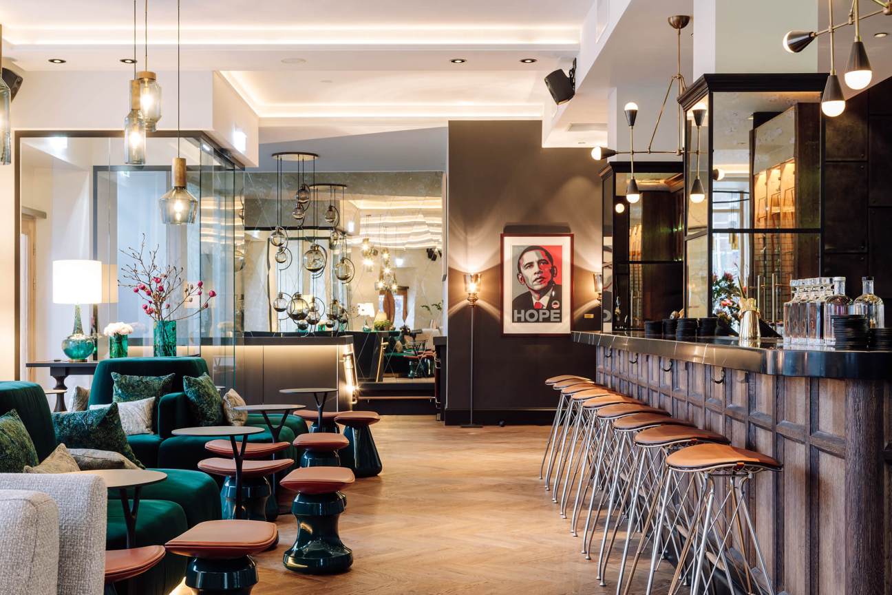 Modern hotel bar with teal seating, wooden counter with bar stools, pendant lighting and a HOPE poster on dark wall.