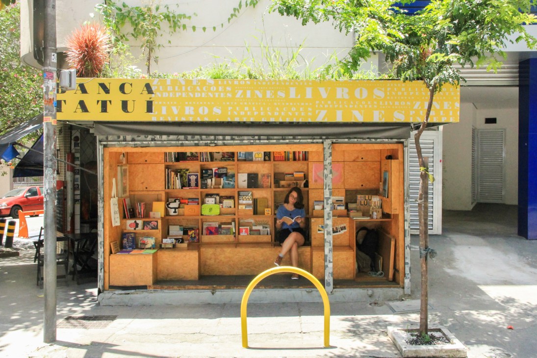 Woman reading inside Banca Tatuí newsstand with orange wooden shelves filled with books and publications.