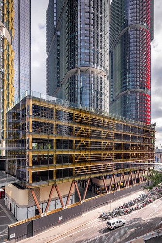 Construction site of International House Sydney showing cross-laminated timber and glass facade with yellow metal framework surrounded by tall city buildings.