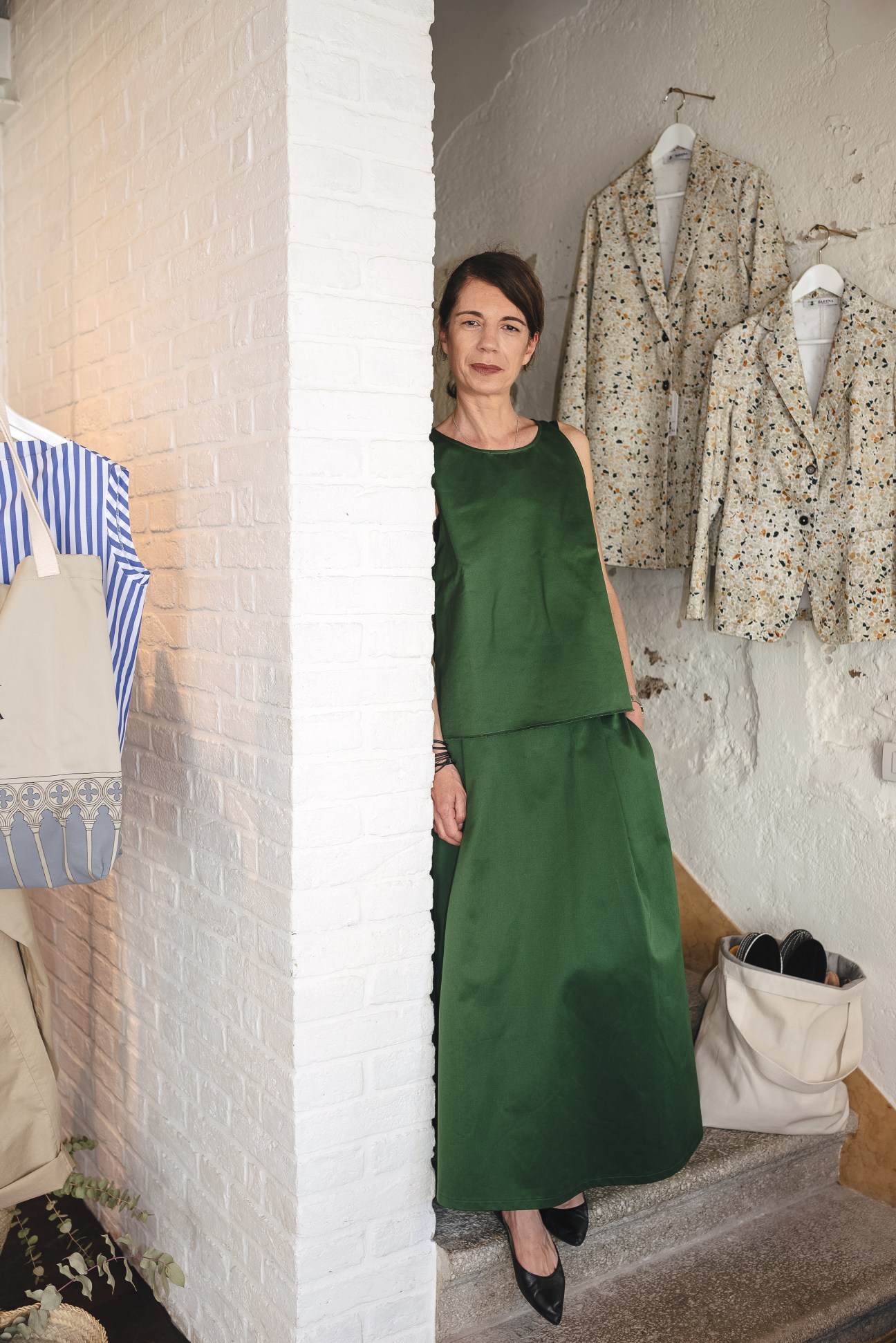 Paola Jani standing in Barena's women's shop wearing a green sleeveless dress with terrazzo-patterned blazers displayed on the wall behind her.