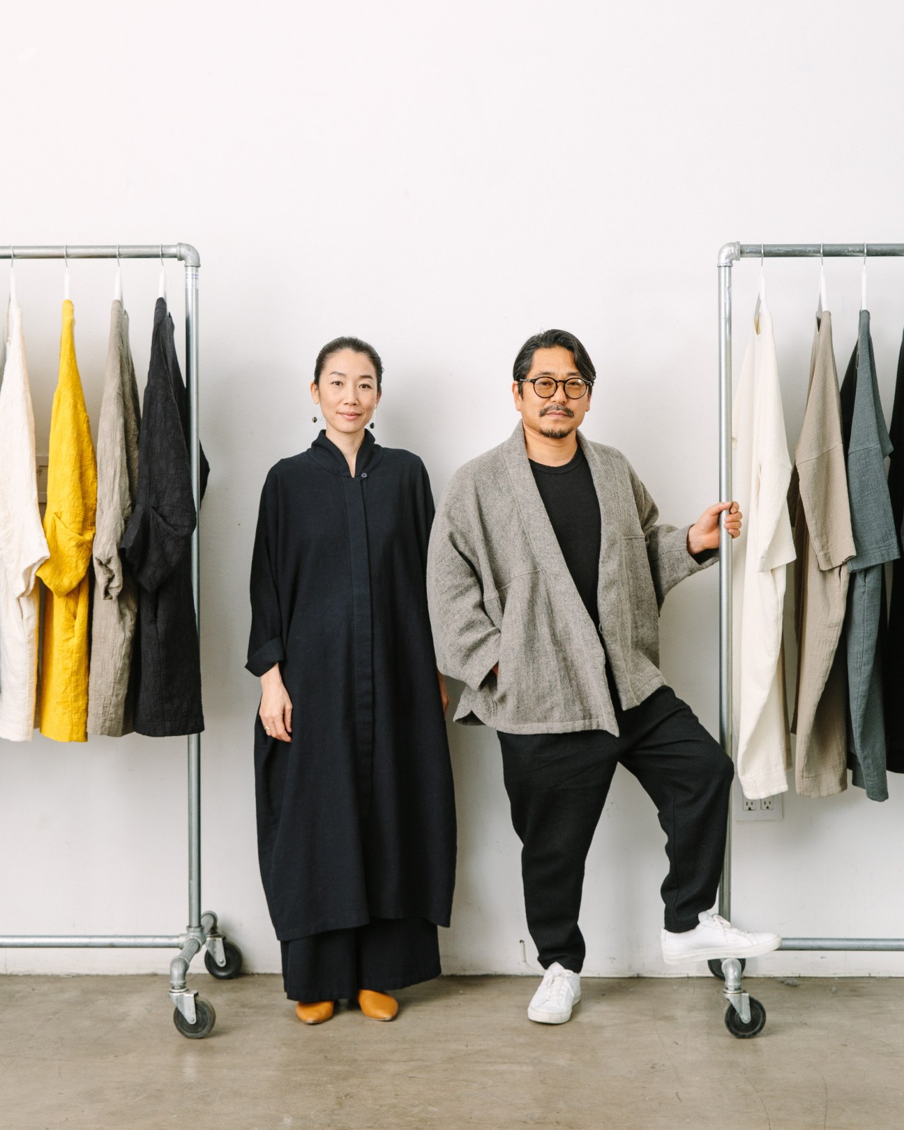 Two designers standing between clothing racks displaying colorful garments in a bright studio space.