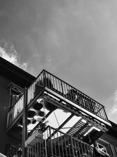 Black and white architectural photograph of a bolt-on balcony attached to a building facade against a cloudy sky.