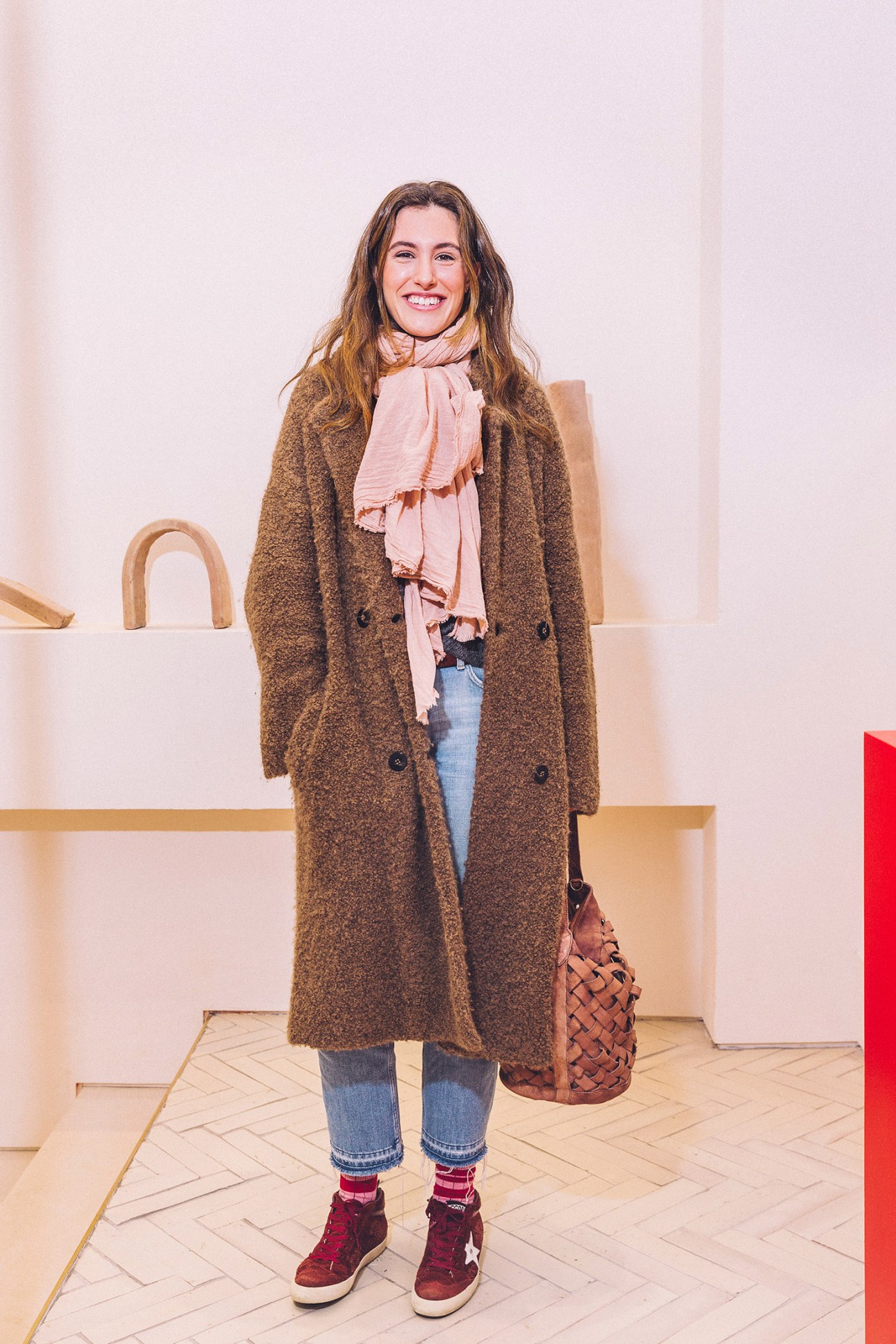 Woman in brown coat and pink scarf holding woven brown bag in modern interior space