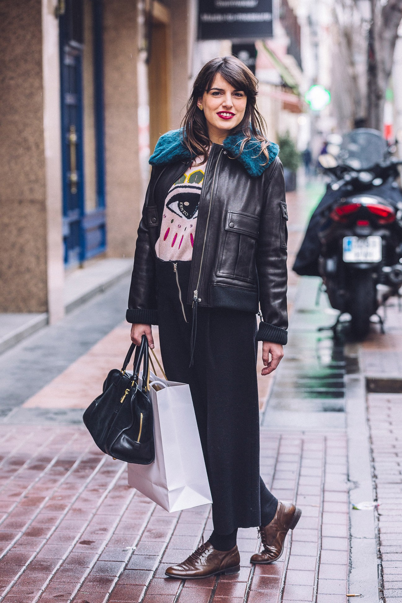 Woman in black leather jacket with teal fur collar holds shopping bag on Madrid street.