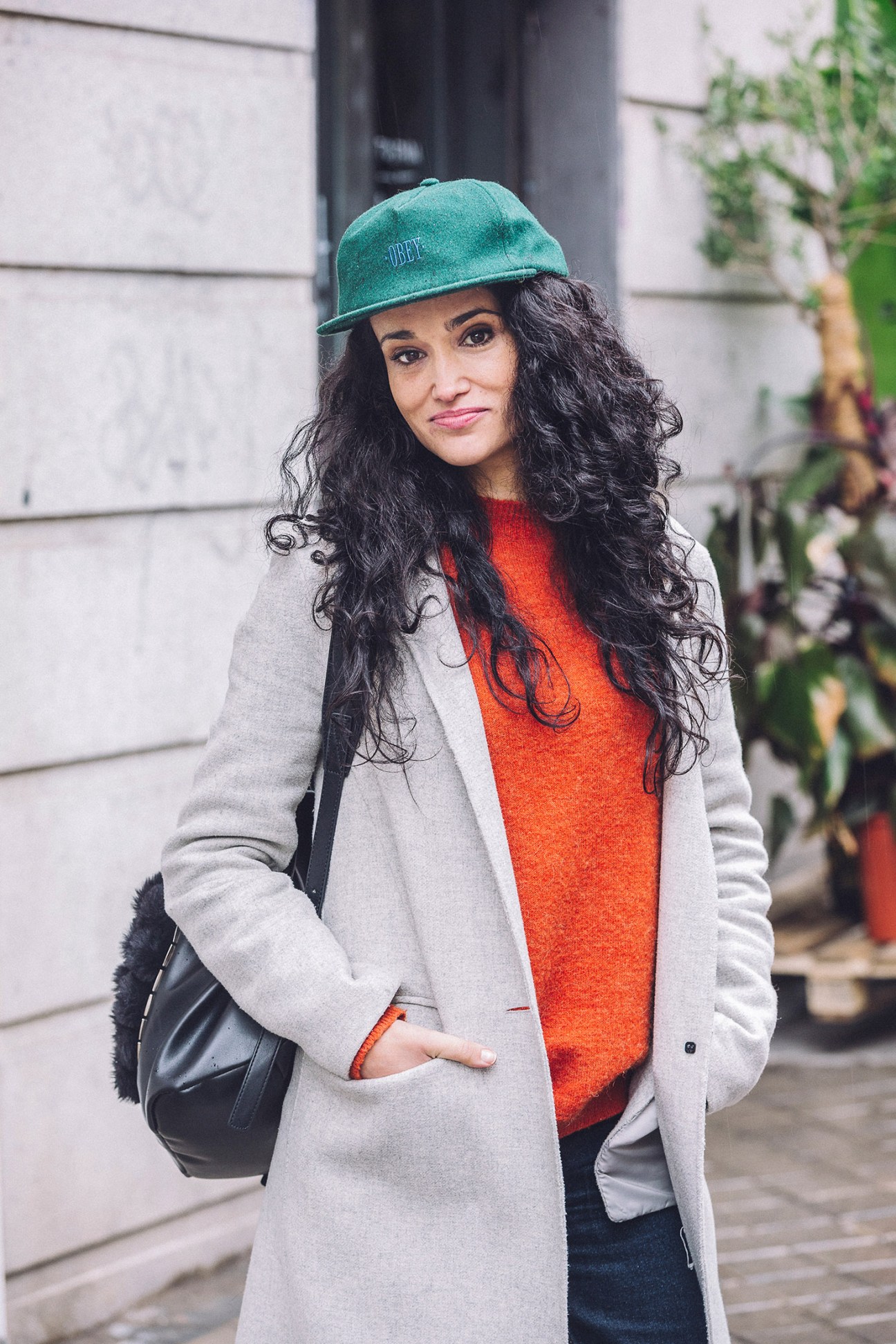 Woman in teal cap, gray coat, and orange sweater poses for street style photo in Madrid.