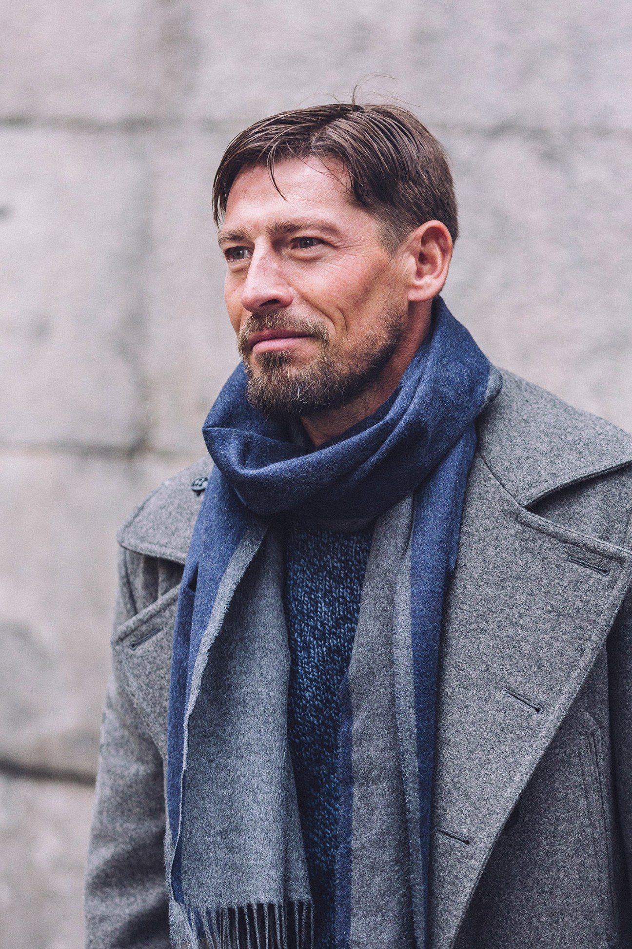 Man with beard wearing grey coat and blue scarf standing outdoors.