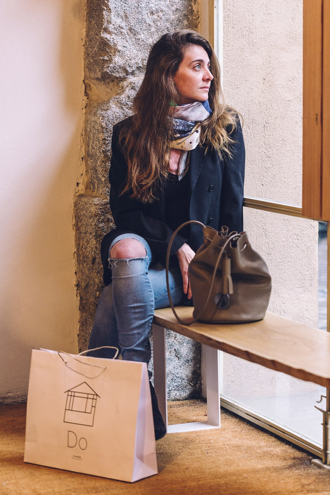 Woman in black blazer and ripped jeans holding brown leather bag sits on windowsill with shopping bag